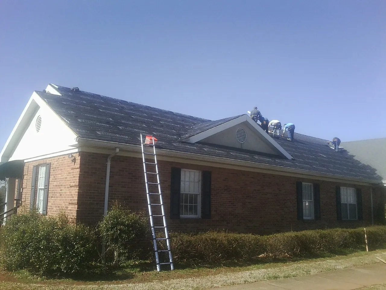 Crew tearing shingles off a brick ranch roof with a ladder set — Emergency Roof Repair work by our crew in Dry Fork