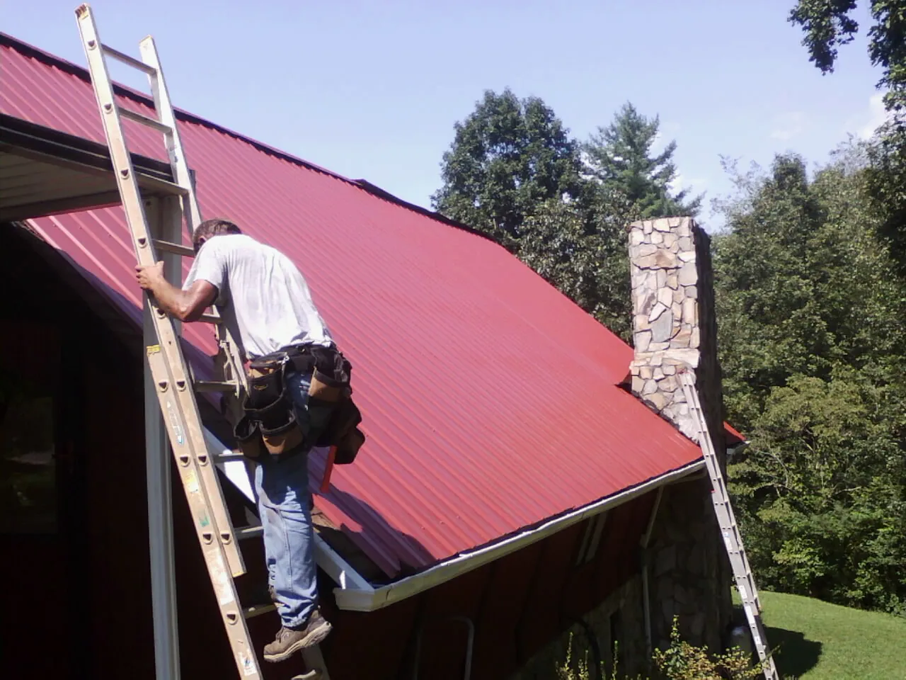 Roofer on a ladder beside a red metal roof and stone chimney — Soffit & Fascia Repair by Roofing Brothers in Dry Fork